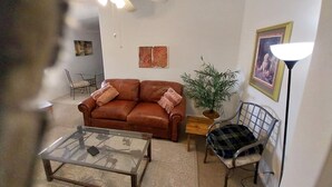 Ein Wohnzimmer mit einem braunen Ledersofa, einem Glastisch, einem Deckenventilator, einer Topfpflanze, einem gerahmten Bild und einer Stehlampe.
