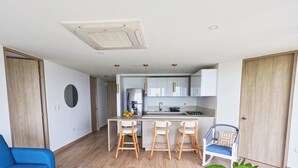 A modern kitchen with white cabinets, a stainless steel refrigerator, and a gas stove. There are two wooden bar stools and a blue chair. - Apartamento familiar en Cartagena, cerca al aeropuerto y playa Las Americas (cartagena de indias)