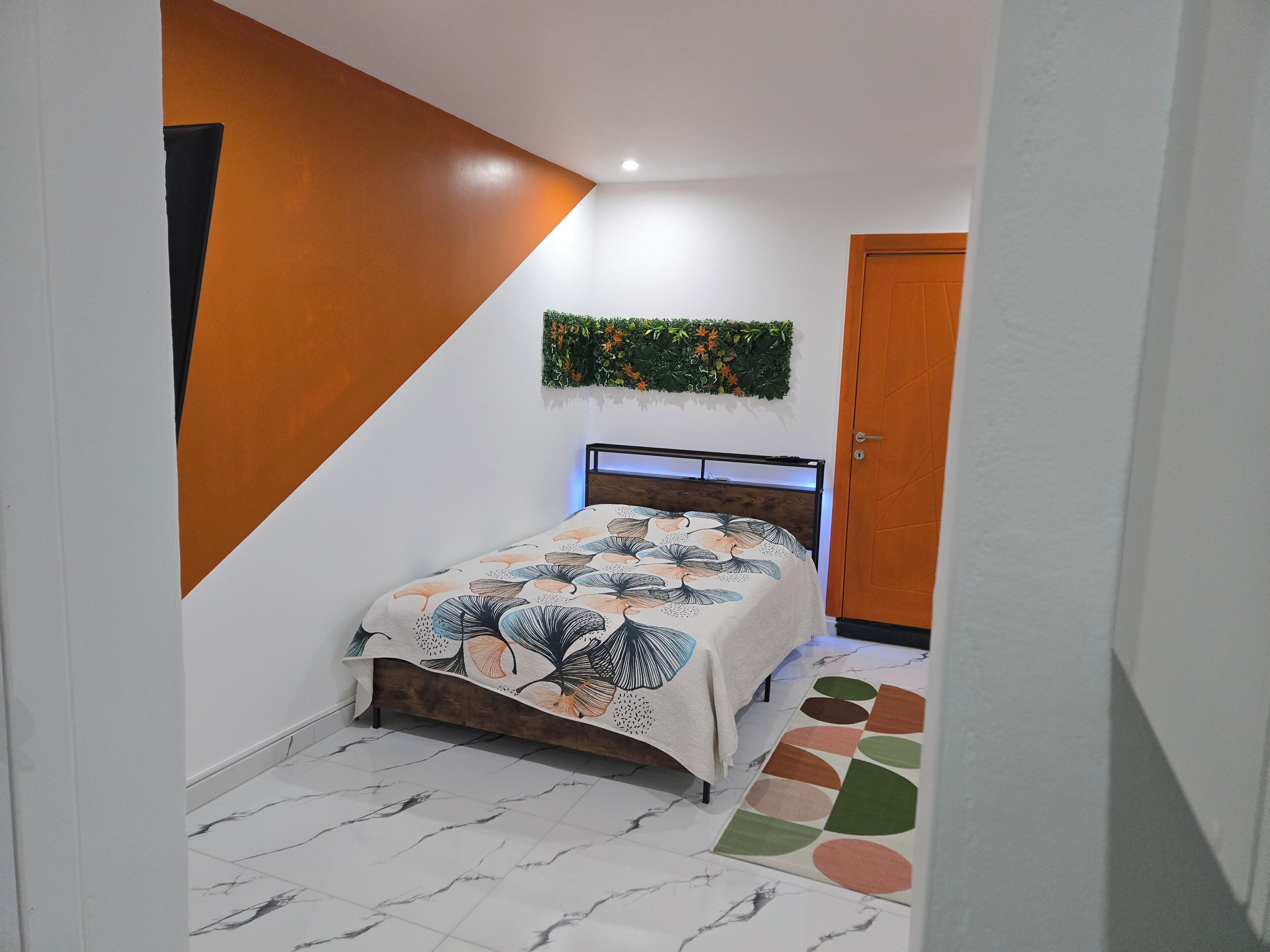 Une chambre à coucher avec un lit, une porte en bois, une plante fixée au mur et un tapis à motifs.