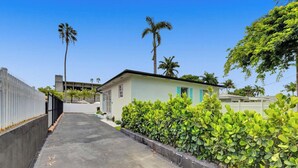 A narrow, paved pathway leading to a light blue building with a flat roof, surrounded by lush green bushes and palm trees. - The Island Shoes (Miami)