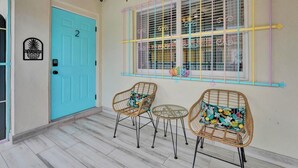 A room with a turquoise door, two wicker chairs with floral cushions, a small round table, and a window with colorful blinds. - The Island Shoes (Miami)