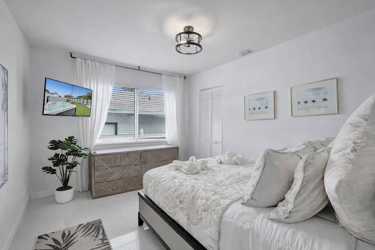 Une chambre à coucher avec un lit, une plante en pot, une fenêtre avec des rideaux, un téléviseur à écran plat et des tableaux encadrés accrochés au mur.