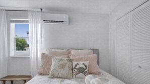 A bedroom with a bed, pillows, a bedside table, an air conditioning unit, and a window with curtains. - The Island Shoes (Miami)
