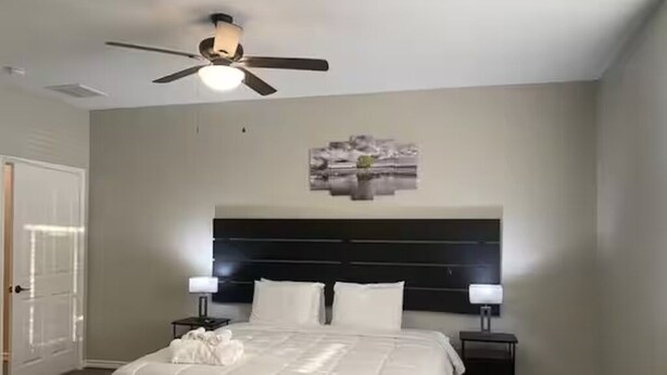 Ein Schlafzimmer mit einem Bett, einem Deckenventilator, einem Nachttisch und einem an der Wand hängenden Kunstwerk.