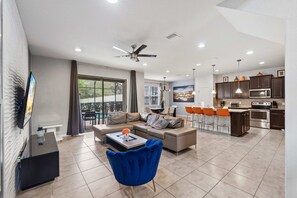 Una sala moderna con sofá, mesa de centro y sillón azul. Se ve una cocina en el fondo con armarios oscuros y electrodomésticos de acero inoxidable.