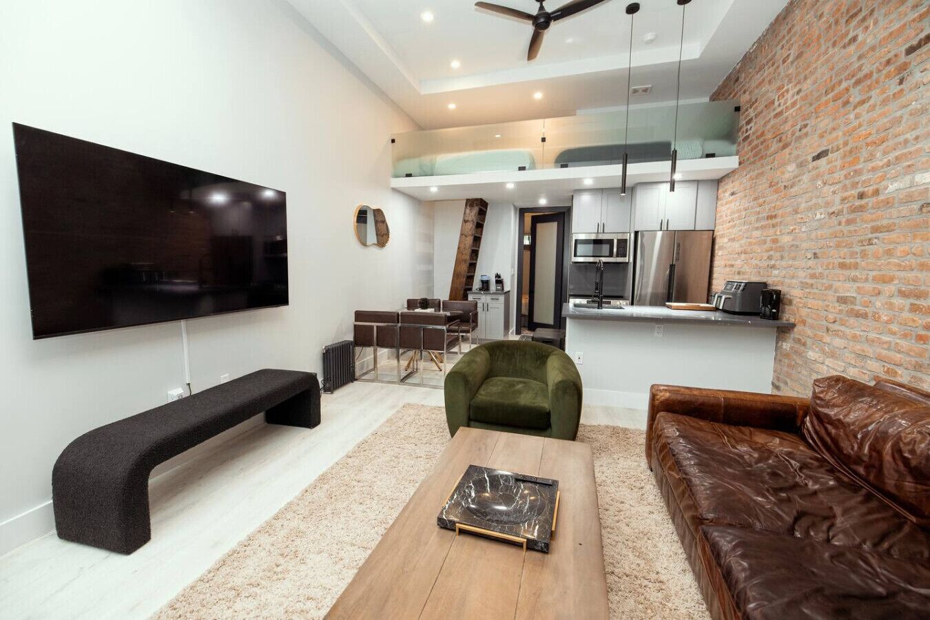 A modern living room with a large flat-screen TV, a wooden coffee table, a green armchair, a brown leather sofa, and a kitchen area in the background.