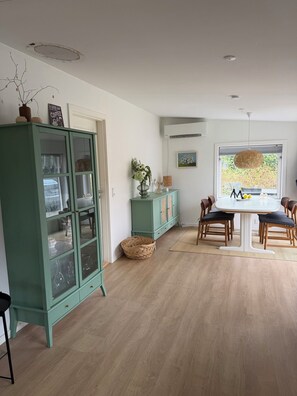 A dining area with a round table, chairs, a woven basket, a cabinet, and a window with a view. - Modernised cottage near woods and beach (Allingåbro)