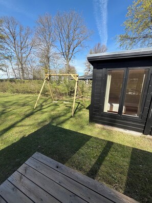 A wooden deck, a black cabin with large windows, and a wooden swing set in a grassy yard. - Modernised cottage near woods and beach (Allingåbro)