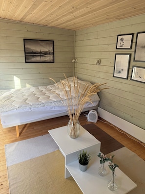 A bedroom with a bed, a bedside table with a plant, a small fan, and a wall-mounted light fixture. - Modernised cottage near woods and beach (Allingåbro)
