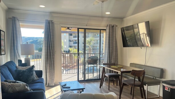 Een moderne woonkamer met een bank, een salontafel, een eettafel met stoelen, een aan de muur bevestigde televisie en een balkon met uitzicht op gebouwen en palmbomen.