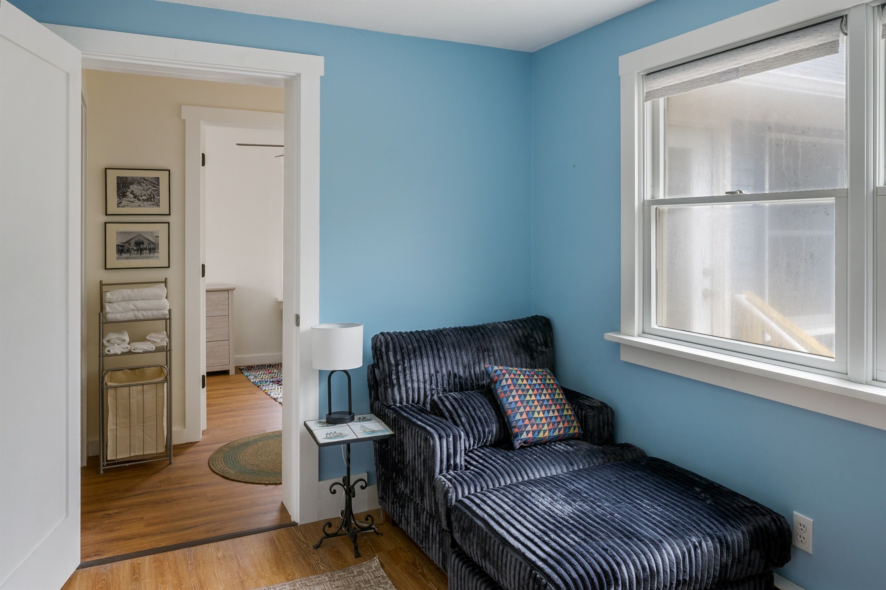 A room with a blue wall, a window, a black sofa with a patterned pillow, a small table with a lamp, and a door leading to another room.