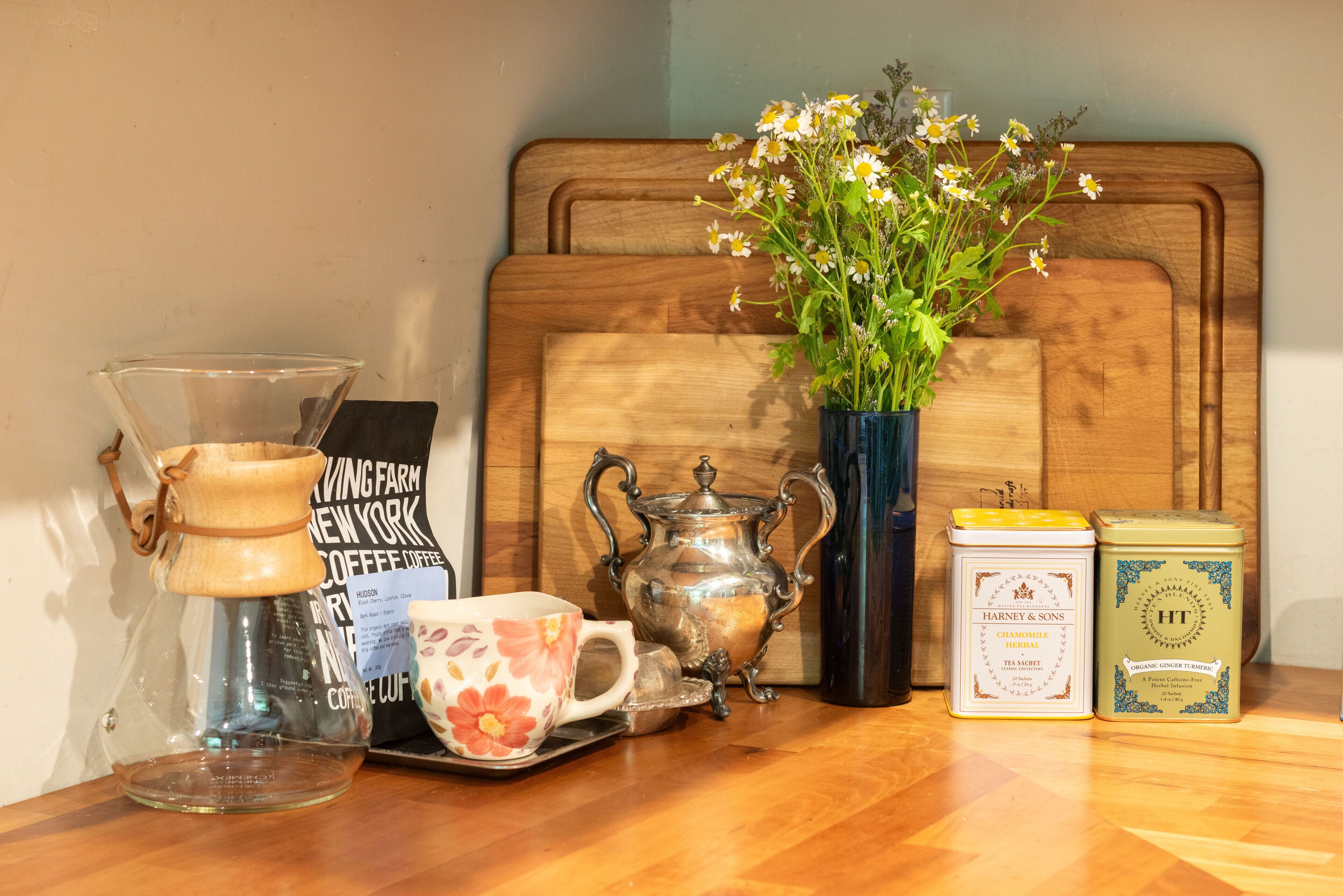 Een houten hoofdbord, een glazen koffiezetapparaat, een mok met bloemen, een vaas met bloemen en twee theedozen.