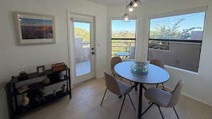 Ein Essbereich mit einem runden Tisch, vier Stühlen, einer Glasbowl, einer schwarzen Konsolentisch mit Dekoration, einem gerahmten Bild und Blick auf einen Balkon mit Holzlatten und Grünpflanzen.
