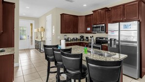 A kitchen with dark wooden cabinets, stainless steel appliances, a granite countertop, and four black bar stools. - 7601 Luxury 7BR Themed Villa w/ Private Pool & Spa (Kissimmee)