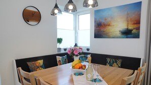 Un coin repas avec une table en bois, des chaises, un saladier de fruits, une bouteille de Prosecco et un tableau représentant un voilier.