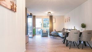 A modern living room with a sofa, dining table, and chairs. There is a large window with curtains, a ceiling light, and a view of the outdoors. - Seestraße 8/13 - Modern. Close to the beach. Sauna. Balcony. (Kellenhusen)