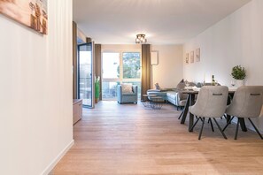Ein modernes Wohnzimmer mit einer Couch, einem Esstisch und Stühlen. Es gibt ein großes Fenster mit Vorhängen, eine Deckenleuchte und einen Blick ins Freie.