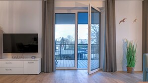 A modern living room with a flat-screen TV on a white cabinet, a sliding glass door to a balcony, and a potted plant. - Seestraße 8/13 - Modern. Close to the beach. Sauna. Balcony. (Kellenhusen)