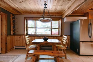 A wooden interior with a dining table, chairs, a hanging light fixture, a window, and a refrigerator. - Cozy King Suite, Perfect Base for Trail & Lake Fun (Cable)