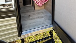 A compact RV interior with a kitchenette, a bed with a red and white blanket, and a window.