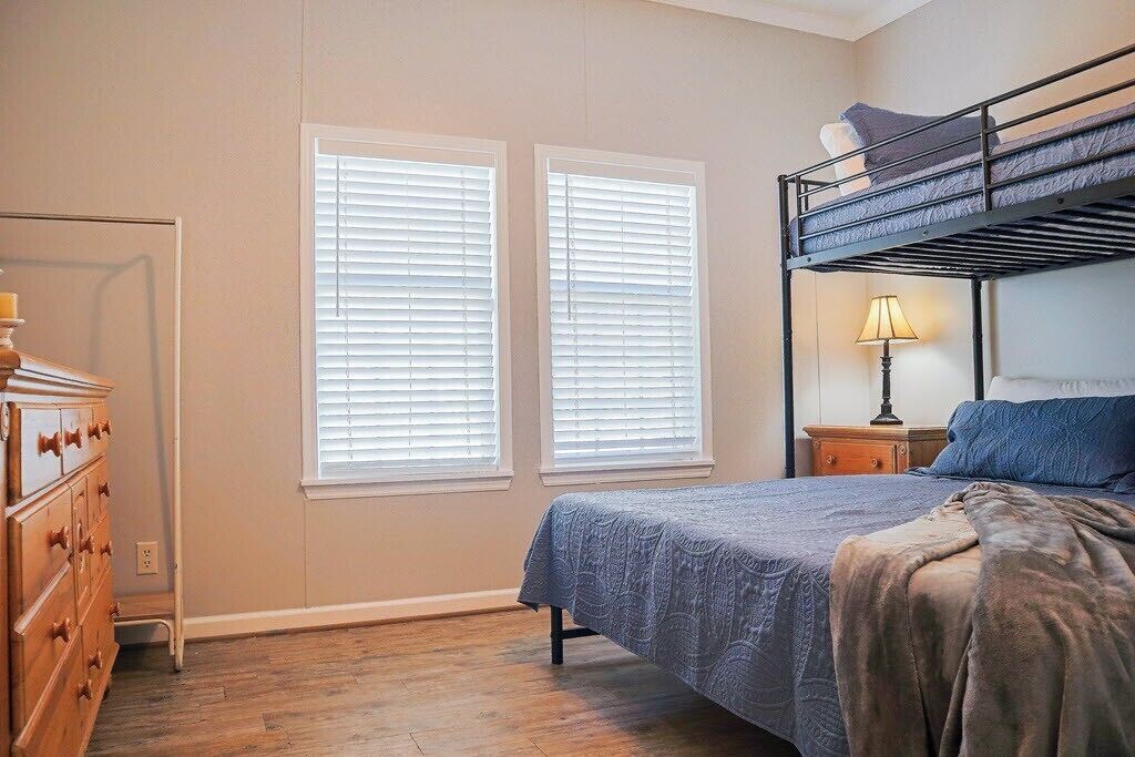 A bedroom with a bunk bed, a dresser, and two windows with blinds.