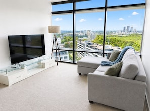 Una sala de estar moderna con un televisor grande, una mesa de centro de vidrio, un sofá gris y vistas a la ciudad a través de amplios ventanales.