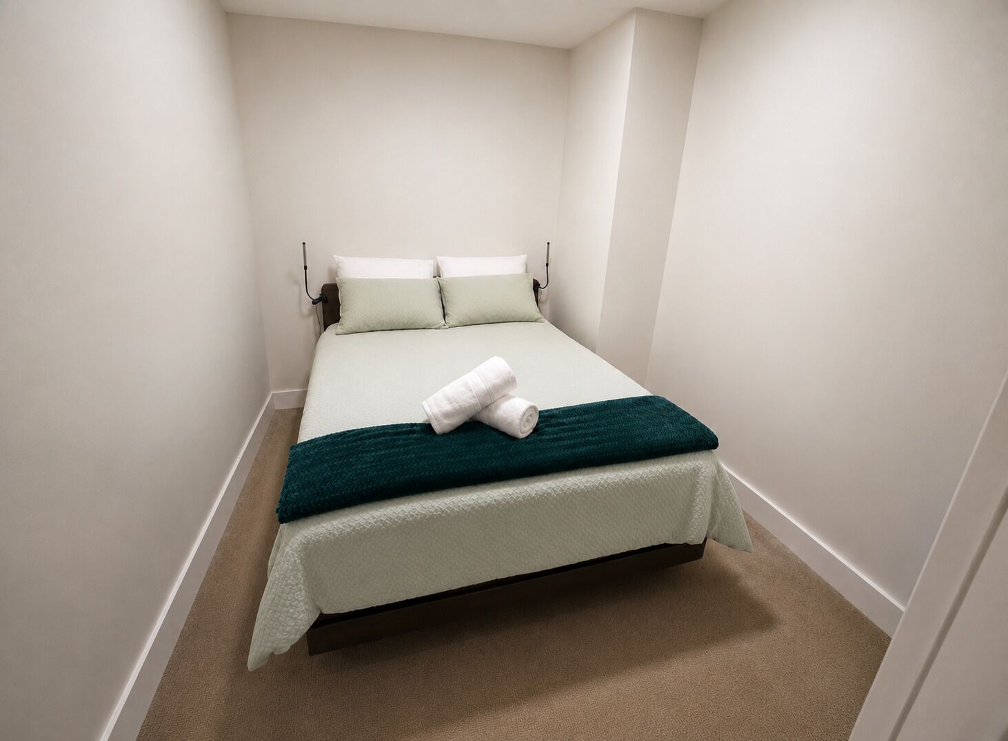 Una cama individual con una manta verde y almohadas blancas en una habitación pequeña de paredes blancas.