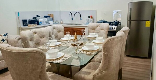 Une salle à manger avec une table en verre dressée pour six personnes, entourée de fauteuils à capitonnage. On aperçoit, au fond, une cuisine équipée d’appareils électroménagers modernes.
