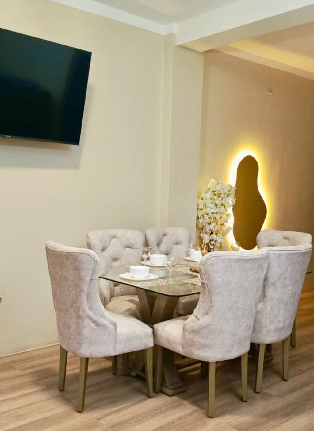 A dining area with a round table, six upholstered chairs, and a wall-mounted TV.