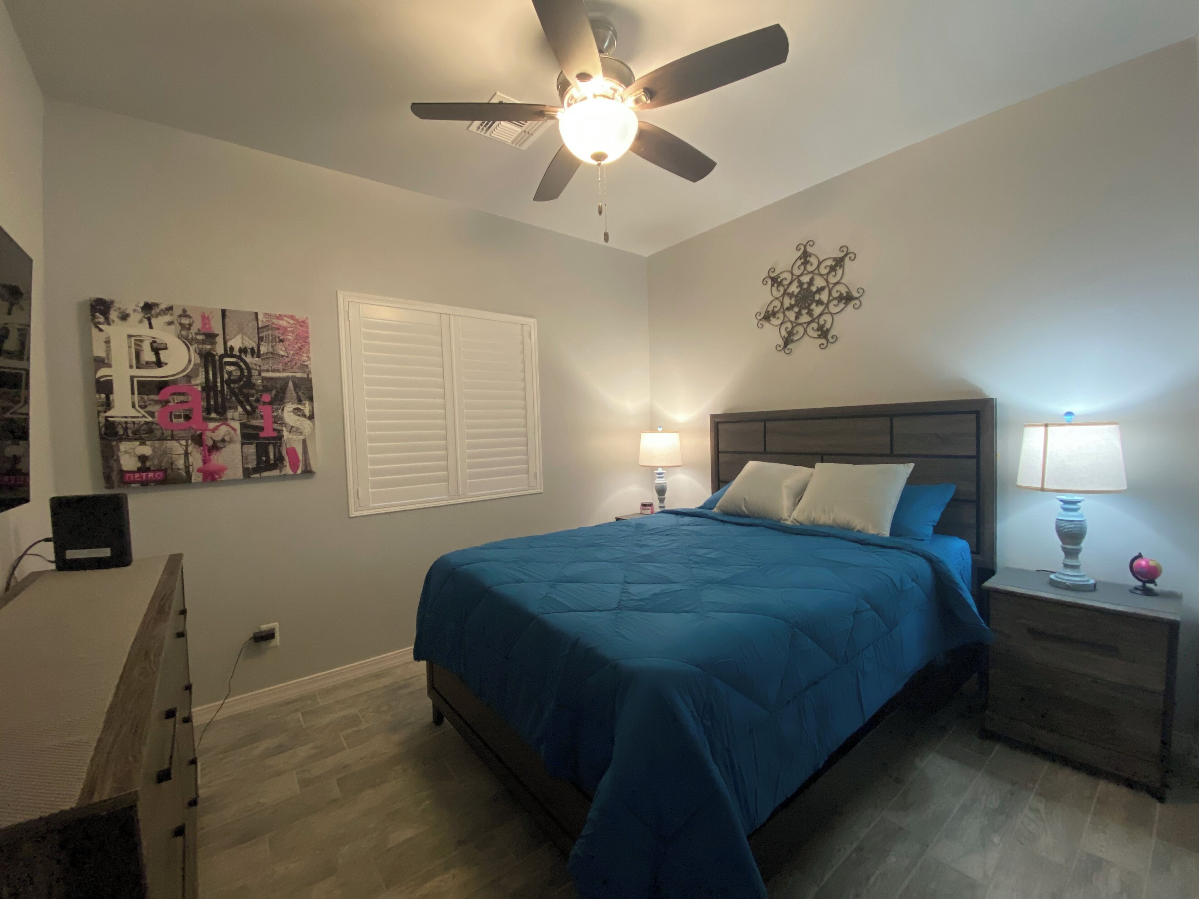 A bedroom with a bed, nightstands, a ceiling fan, a wall-mounted air conditioning unit, and a decorative wall piece.