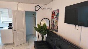Una sala de estar con un sofá de cuero negro, un televisor montado en la pared y una puerta que da a una cocina.