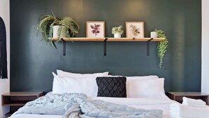 A neatly made bed with white linens and a gray blanket, flanked by two bedside tables with white lamps. Above the bed, there are three wooden shelves holding various plants and framed botanical prints.