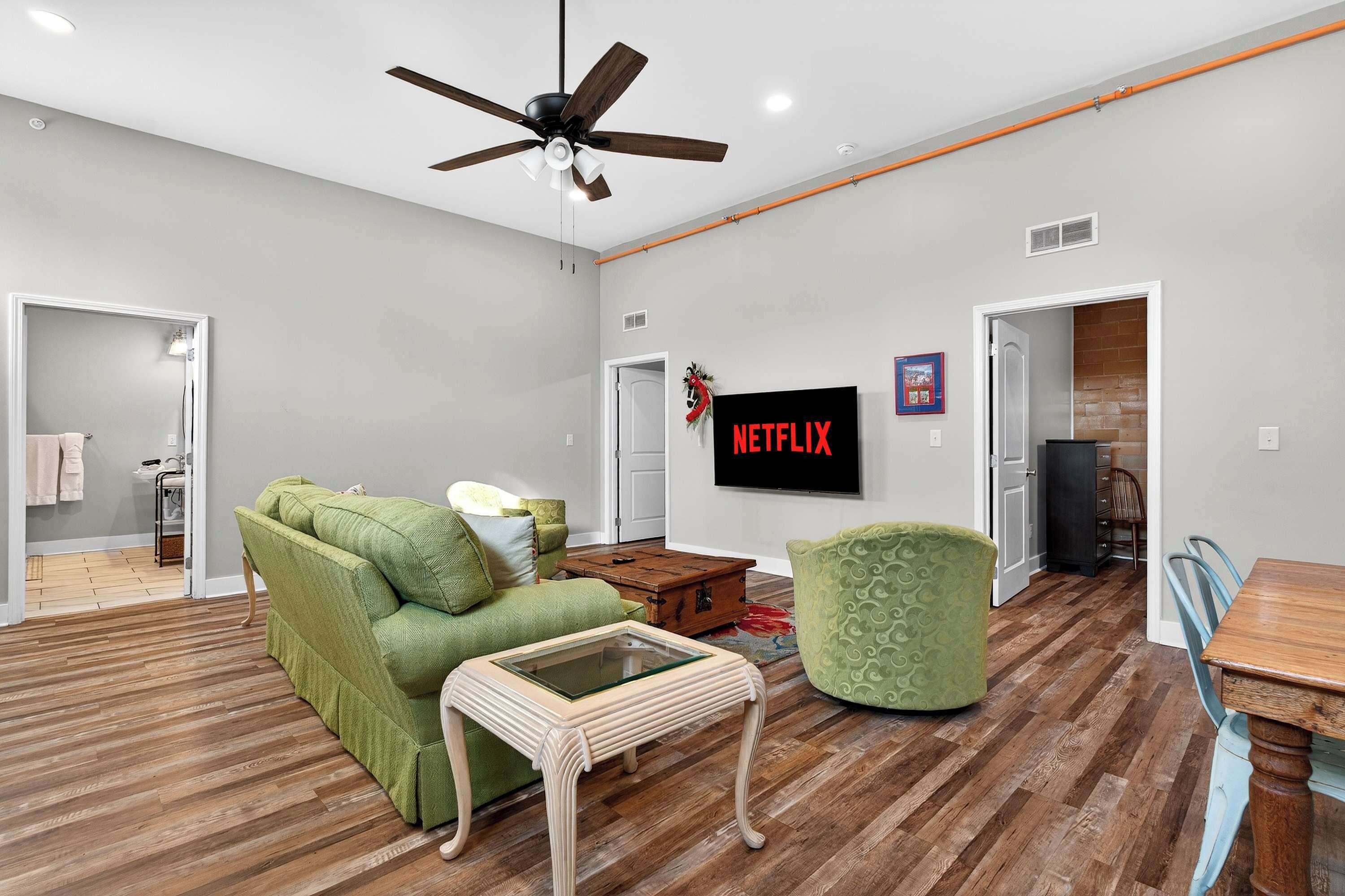 A spacious living room with a green sofa, a wooden coffee table, a ceiling fan, and a flat-screen TV mounted on the wall.