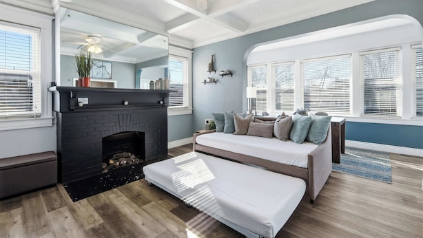 A modern living room with a fireplace, a sofa, and a large window with blinds. - Charming Historic Foursquare Home | 4BR | Backyard (Louisville)