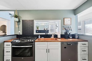 Une cuisine équipée d’une cuisinière à gaz, d’un four, de meubles blancs, d’un plan de travail en bois, d’un crédence noire et d’une fenêtre avec des stores.
