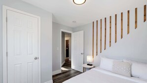 A hotel room with a large bed, a bedside table with a lamp, a wooden wall panel, and a door leading to another room. - Charming Historic Foursquare Home | 4BR | Backyard (Louisville)