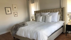 Una cama grande con cabecera tapizada, ropa de cama blanca y varios almohadones. Se observan cuadros enmarcados en la pared y una ventana con cortinas.