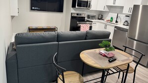 A modern kitchen with white cabinets, a grey sofa, a dining table with chairs, and a flat-screen TV mounted on the wall. - Alake Villa (Saskatoon)