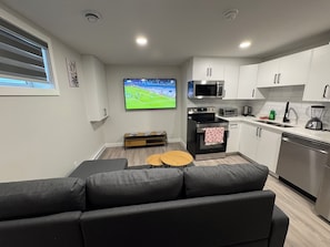 A modern kitchen with a built-in oven, microwave, and dishwasher. There is a small dining table and a grey sofa in the living area. - Alake Villa (Saskatoon)