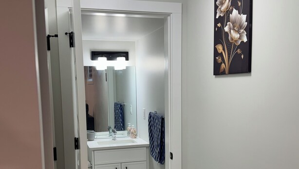A bathroom with a white sink, mirror, and cabinets. There is a towel hanging on the wall and a small rug on the floor. - Alake Villa (Saskatoon)