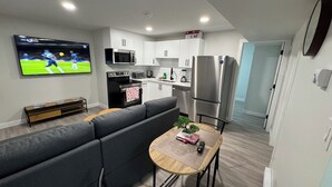A modern kitchen with white cabinets, a stainless steel refrigerator, and a microwave. There is a TV mounted on the wall displaying a sports game. - Alake Villa (Saskatoon)