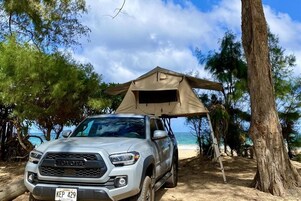 A Toyota SUV parked under a tent on a beach with trees and ocean in the background.