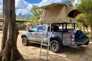 Una camioneta pickup plateada con una tienda de campaña en el techo, estacionada en un entorno natural con árboles y montañas al fondo.