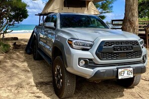Uma caminhonete Toyota prata estacionada em uma praia arenosa, com uma tenda ao fundo.
