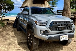 Una camioneta Toyota plateada estacionada en una playa arenosa con una tienda de campaña al fondo.