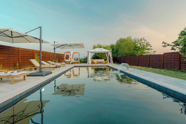 A swimming pool with lounge chairs and umbrellas, surrounded by a wooden fence and greenery.