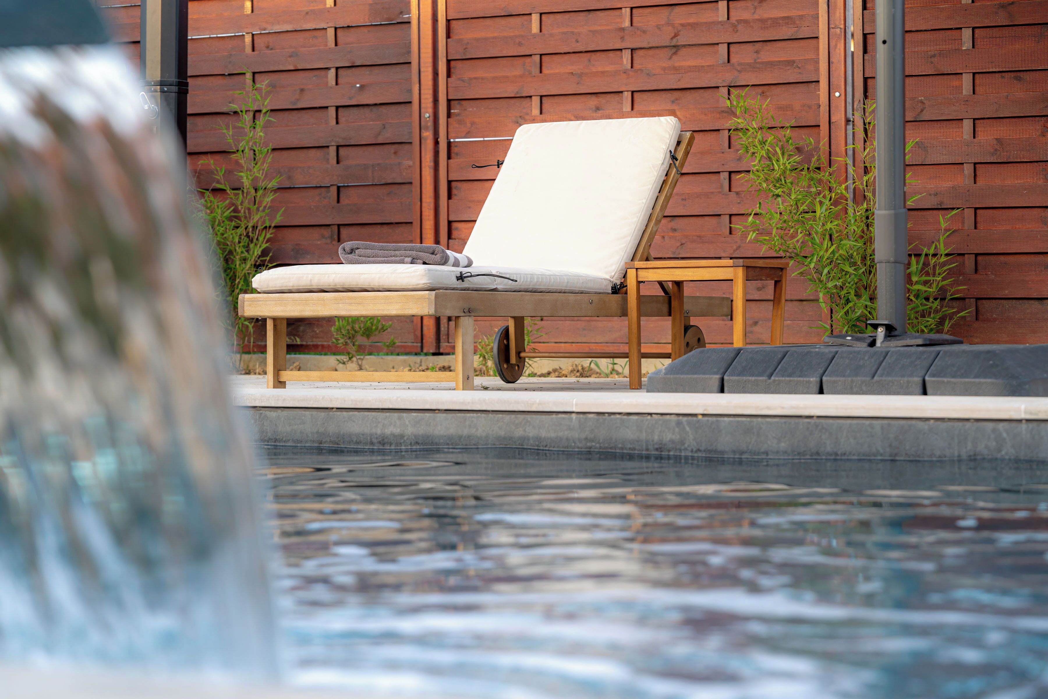 Una silla de lounge de madera con un cojín y una toalla, ubicada junto a una piscina con una valla de madera al fondo.