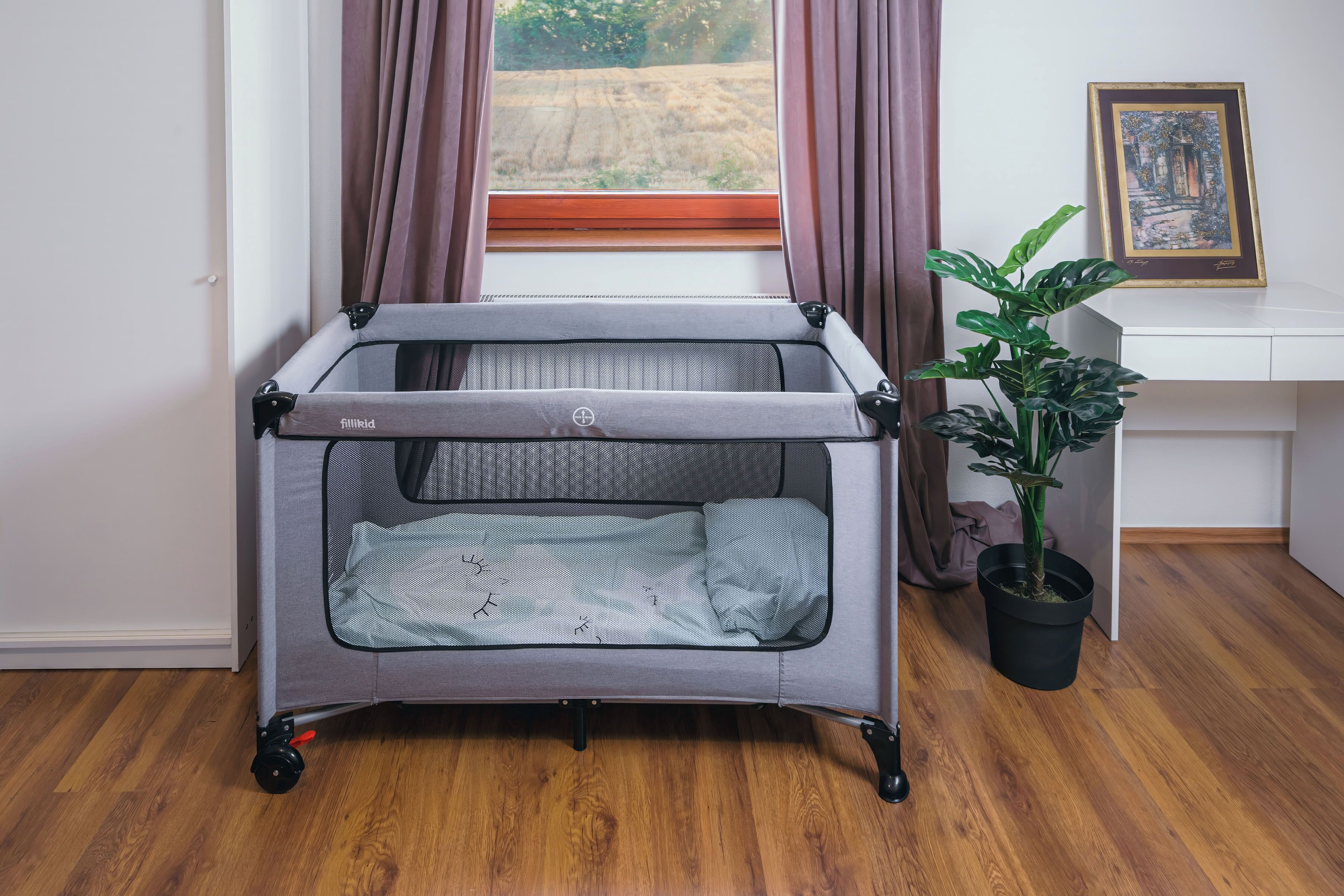A room with a grey portable crib, a potted plant, a framed picture on the wall, and a window with curtains.