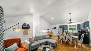 A spacious living area with a gray sofa, a red armchair, and a dining table with patterned chairs. A kitchen with blue cabinets and a hanging light fixture is visible in the background. - Little Toasty / 9 min to Dollywood! 8 min to Pkwy! (Pigeon Forge)