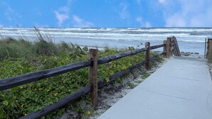 A wooden fence leading to a beach with waves crashing onto the shore. - Monthly Coastal Retreat | Garage, Pools, Beach Access & Quiet Community (New Smyrna Beach)